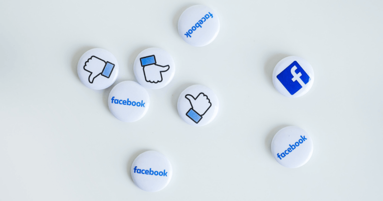 Na białym stole przypinki z logotypem Facebooka, nazwą Facebook i znakiem lajka. Jest ich 8, ułożone w estetycznym nieładzie.