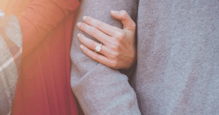 Na zdjęciu dwie osoby, zbliżenie na ich splecione ręce. Na środku dłoń z pierścionkiem zaręczynowym. Jedna postać ma czerwony sweter prążkowany, druga szary, gładki.